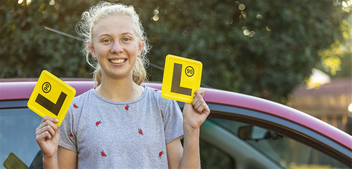 A smiling young female wearing a grey t-shirt is standing next to a red car, and holding 2 yellow L Plates in her hands