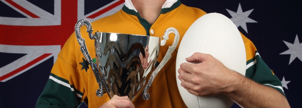 A man dressed in yellow and gold jersey holding a trophy in one hand and a ball in the other. An Australian flag is in the background.