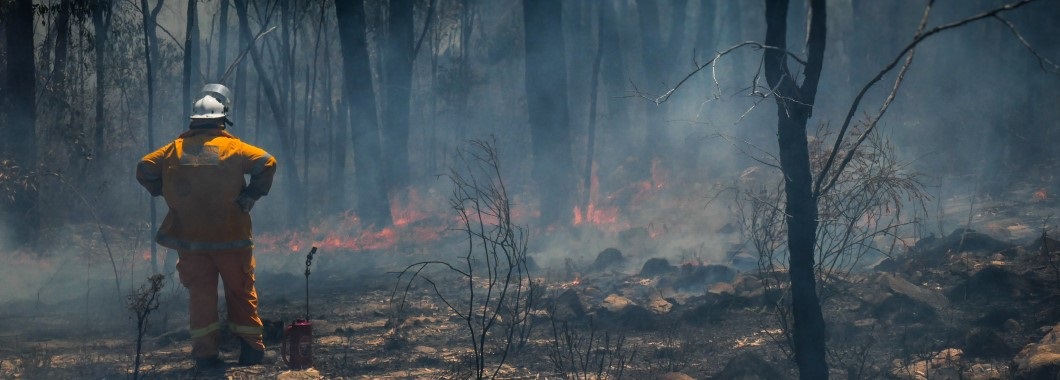 Fire fighter watching over burnt bush land with back burning tool, small flames burning in the background