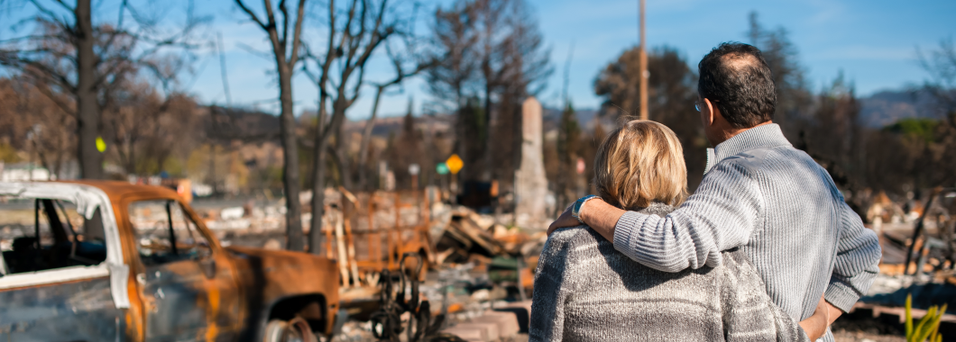 Mature aged couple holding each other looking at fire damage including a burnt car, tree and lost homes