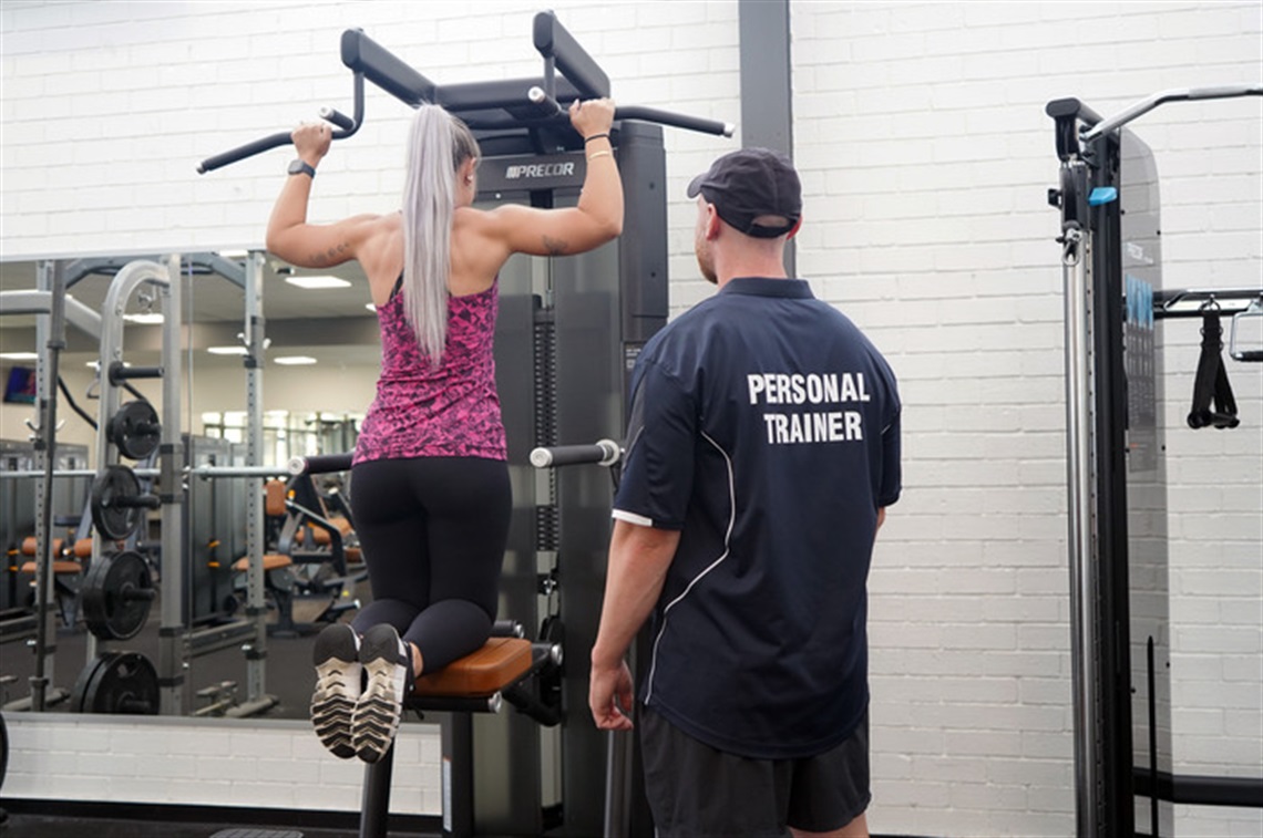 Customer using gym equipment with personal trainer watching